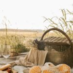 Tranquil picnic scene with wicker basket, wine, oranges, and bread on a grassy countryside field.